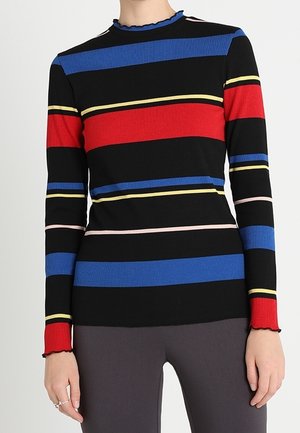 Nainen pukeutuneena mustaan tiukasti istuvaan ribbipuseroon, jossa on vaakasuorat raidat punaisessa, sinisessä, keltaisessa ja pinkissä, yhdistettynä tummanharmaisiin housuihin.