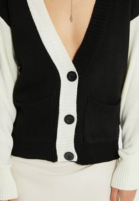 Czarnobiały dziergany kardigan z głębokim dekoltem w szpic, dwoma przednimi kieszeniami i czarnymi guzikami wzdłuż środka. Luźny krój z prążkowaną fakturą.