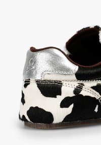 Zapatilla con estampado de vaca que presenta una textura suave similar a la piel, acento metálico plateado en el talón y forro de ante marrón con detalles de costuras visibles.