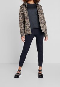 Kvinna som bär en kamouflagejacka, mörk långärmad tröja, svarta träningsleggings och svarta sandaler med vita sulor, stående på en grå golv.