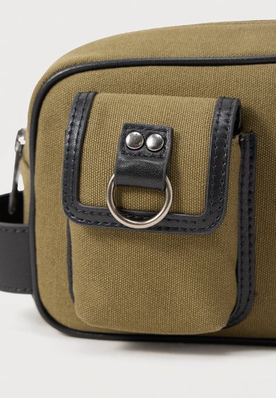 Μικρό υφασμάτινο πουγκί ελιάς με μαύρο δερμάτινο περίγραμμα, κλειστό με καπάκι με ασημένιες τρουκ και στρογγυλό μεταλλικό δαχτυλίδι στο μπροστινό μέρος.