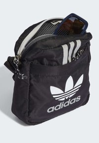 Borsa a tracolla nera Adidas con logo bianco, parzialmente zipata, contenente occhiali da sole con montatura blu all'interno del compartimento principale.