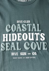 Grön bomullströja med upphöjd vit och ljusblå text: "DIVE CLUB COASTAL HIDEOUTS SEAL COVE DIVE SUN and CO. DAILY RAYS and DEEP DIVING."