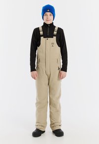 Beige sneeuwoverall met schouderbanden, met ritssluiting afgesloten zakken en een zwart longsleeve shirt eronder. Draag het met een blauwe muts en zwarte laarzen.