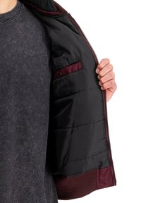 Padded schwarzer Mantel mit einem burgunderroten Futter, ausgestattet mit einem Reißverschluss und gerippten Bündchen. Das Innenfutter umfasst eine kleine Tasche mit einem Knopf-Detail.