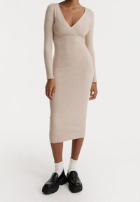 Abito lungo a maniche lunghe beige a coste con scollatura a V e silhouette aderente, abbinato a mocassini neri con plateau e calze bianche a misura.