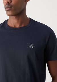 Mornaricki pamučni T-shirt s okruglim izrezom. Na prsima se nalazi bijeli CK logo i natpis "Calvin Klein Jeans". Kratki rukavi, glatka tekstura.