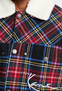 Rutigt skjorta med röda, gröna, blå och gula mönster. Har en vit fleecekrage och silverfärgade tryckknappar för stängning.