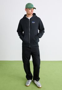 Young man in black zip-up hoodie, black cargo pants, silver sneakers, and green cap standing on green floor against white wall.