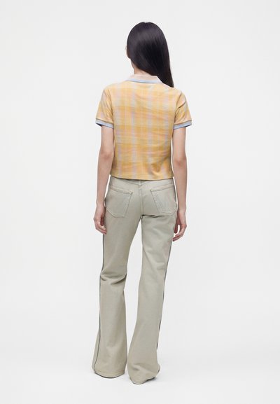 Femme debout de dos portant une chemise à carreaux jaune à manches courtes et un pantalon évasé beige clair avec des poches arrière sur fond blanc.