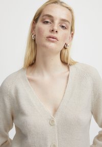 Beige knitted cardigan with a V-neck, featuring a button closure and ribbed edges. Smooth texture and lightweight material.