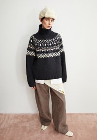 Schwarzer Pullover mit hohem Kragen, versehen mit einem weißen geometrischen Muster. Kombiniert mit braunen, weit geschnittenen Wollhosen und einem cremefarbenen Strickhut.