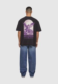 Svart oversized t-skjorte med et lilla grafisk motiv av en vinget figur med et spyd på ryggen. Løse blå denimbukser fullfører stilen.
