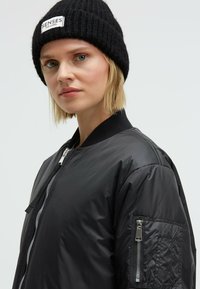 Black bomber jacket with a smooth, shiny texture and zippered pockets. Paired with a black knitted beanie featuring a label.