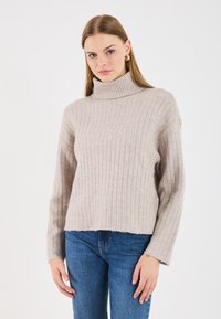 Pulover turtleneck de culoare bej deschis, cu mâneci lungi, textură moale, croială lejeră și tiv scurt, asortat cu blugi albaștri.
