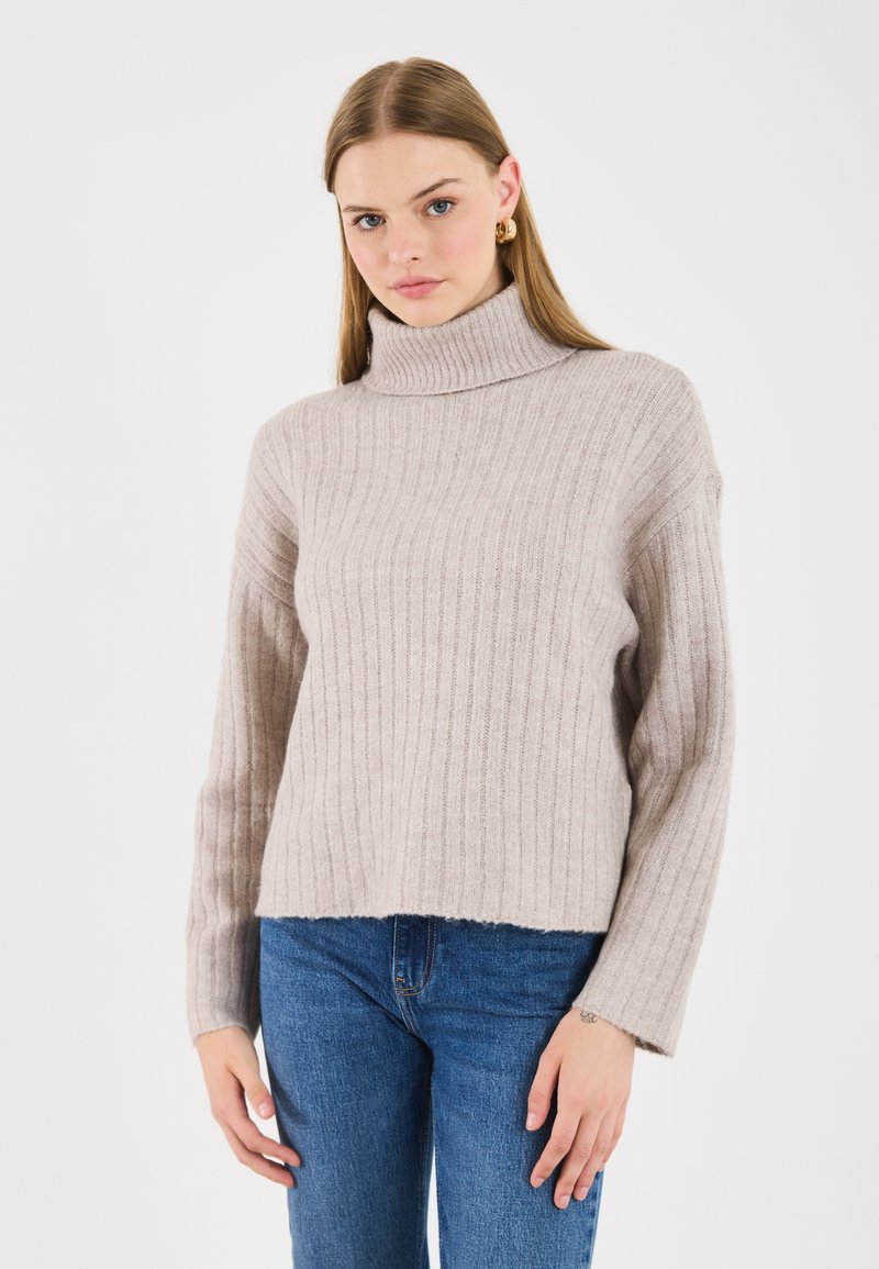 Hellbeige, gerippter Rollkragenpullover mit langen Ärmeln, weichem Textur, lockerer Passform und abgerundetem Saum, kombiniert mit blauen Jeans.