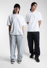 Twee personen dragen oversized, eenvoudige witte T-shirts. De één draagt ook grijze joggingbroeken en de ander donker, baggy jeans met zwarte schoenen.