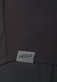 Čierne športové tričko s hladkým povrchom, s logom značky HOKA v šedej farbe a pruhmi umiestnenými na leme. Jednoduchý a funkčný dizajn.
