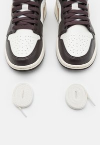 Sneakers in pelle marrone e bianca con punta rotonda, parte superiore perforata e lacci piatti. Include due paia extra di lacci bianchi accanto alle scarpe.