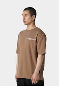 Brun oversized t-skjorte med korte ermer, med et hvitt logo-print på venstre bryst og en avslappet passform. Glatt tekstur og enkelt design.