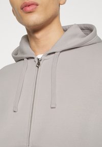 Gros plan sur une personne portant un sweat à capuche gris clair avec fermeture éclair et cordons, partiellement ouvert pour révéler un t-shirt blanc en dessous.