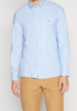 Mand iført en lyseblå langærmet skjorte med knapper og beige bukser, stående foran en ensfarvet lys baggrund.