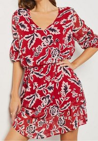 Vestido floral rojo con escote en V, mangas abullonadas y cintura ceñida. La tela es transparente con un dobladillo en capas, presentando intrincados patrones oscuros y claros.