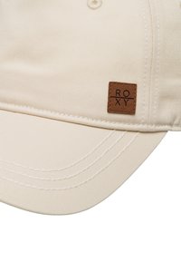 Gorra de béisbol color crema fabricada con tejido duradero, con una superficie texturizada, costuras bordadas y un parche de logo de cuero marrón.