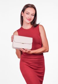 Světlý béžový psaníčko s klopovém uzavřením, zlatými detaily, obdélníkovým tvarem a hladkým povrchem, které drží osoba v červených šatech.