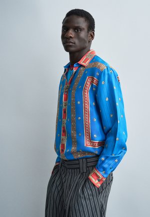 Νέος άντρας που φοράει μια ζωντανή μπλε πουκάμισο με κόκκινες και χρυσές λεπτομέρειες και ασπρόμαυρη ριγέ παντελόνι, στέκεται μπροστά σε ένα απλό φόντο.