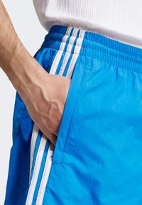 Bermudas azuis com três riscas brancas ao longo dos lados, cintura elástica e um bolso lateral. O tecido parece leve e suave.