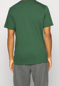 Osoba v jednoduchém zeleném tričku s krátkým rukávem a šedých kalhotách, zobrazená zezadu na světlém pozadí.