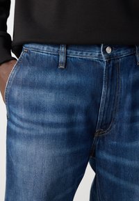Man in blauwe denim jeans met een hand in de zak en een zwart vest dat in de taille is gestopt.