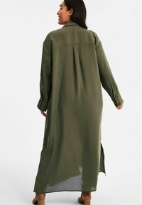 Langes olivengrünes Hemdkleid mit lockerer Passform, geknöpften Manschetten und seitlichen Schlitzen, gefertigt aus einem leichten, glatten Stoff.