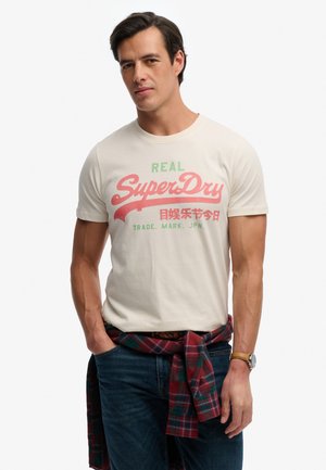 Mann trägt off-white "SuperDry" Logo-T-Shirt, mit kariertem Hemd um die Taille gebunden, blaue Jeans und Armbanduhr, steht mit den Händen in den Taschen.