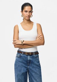 Γυναίκα με σκούρα μαλλιά μαζεμένα πίσω, που φοράει χρυσούς κρίκους στα αυτιά, λευκό αμάνικο τοπ, μπλε τζιν με καφέ ζώνη και χρυσό βραχιόλι, με σταυρωμένα τα χέρια.