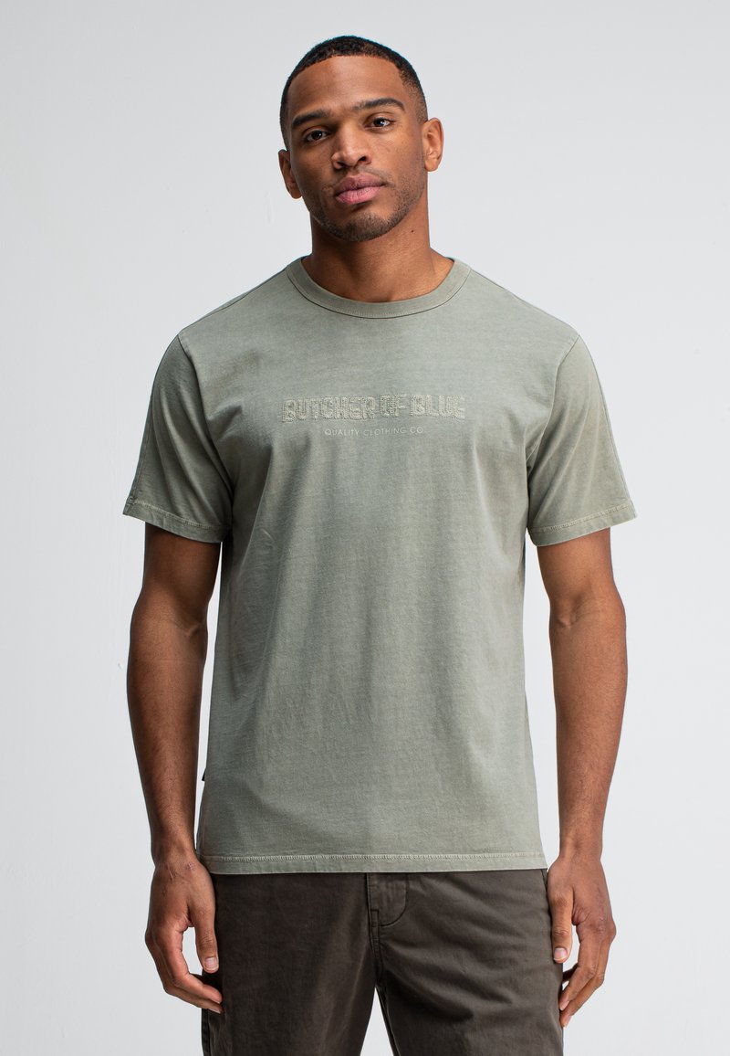 Man in een vervaagde groene "Butcher of Blue" t-shirt en donkere broek, staande tegen een effen lichte achtergrond, kijkend naar de camera.