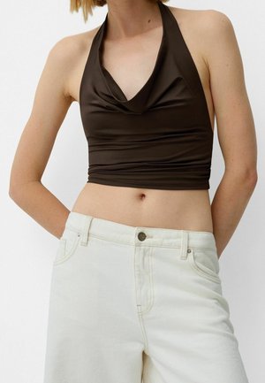 Femme portant un haut marron foncé sans manches à col halter, associé à un jean taille haute couleur ivoire, raccourci au niveau du ventre, debout devant un fond uni.