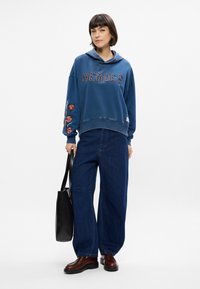 Μπλε υπερμεγέθη φούτερ με κείμενο "HEROES", λουλουδάτα κεντήματα στα μανίκια, συνδυασμένο με φαρδιά μπλε τζιν παντελόνι, καφέ παπούτσια και μαύρη τσάντα.