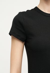Gros plan sur une personne portant un t-shirt noir ajusté à manches courtes et col rond, montrant la partie supérieure du bras et l'épaule.
