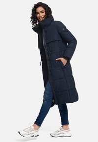 Marinblå lång pufferjacka med hög krage, frontdragkedja, två sidofickor och dragsko i midjan. Bärs över en grå topp och jeans.
