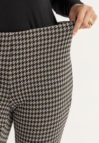 Persoon in zwart en beige houndstooth-patroon broek trekt de tailleband naar buiten om de rekbaarheid te laten zien, hand met gouden ring zichtbaar.