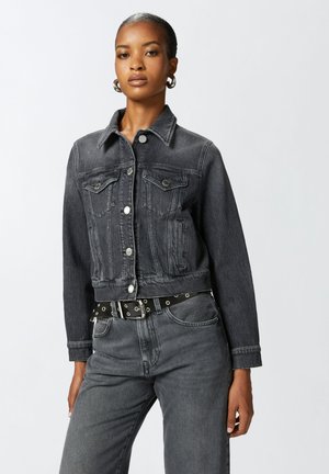 Giacca cropped in denim nero con colletto classico, tasche frontali, chiusura con bottoni a scatto, abbinata a jeans neri con cintura, design minimalista.
