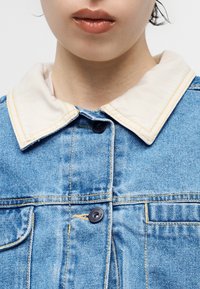 Κοντινό πλάνο ενός ατόμου που φοράει μπλε τζιν μπουφάν με λευκό γιακά αντίθεσης και μαύρα κουμπιά, φαίνονται τα χείλη αλλά όχι άλλα χαρακτηριστικά του προσώπου.