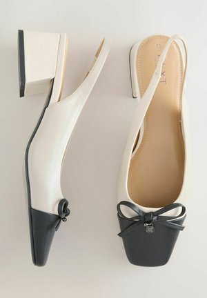 Slingback topánky s blokovým podpätkom z krémovej kože s čiernou špičkou, zdobené dekoratívnou mašľou a malým príveskom na prednej strane.