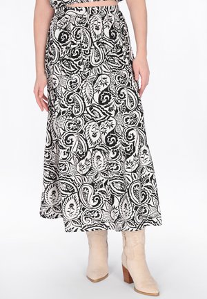 Schwarz-weißer Paisley-Muster-Midirock, getragen mit beigefarbenen, Absatzstiefeln, an einer Person stehend vor weißem Hintergrund.