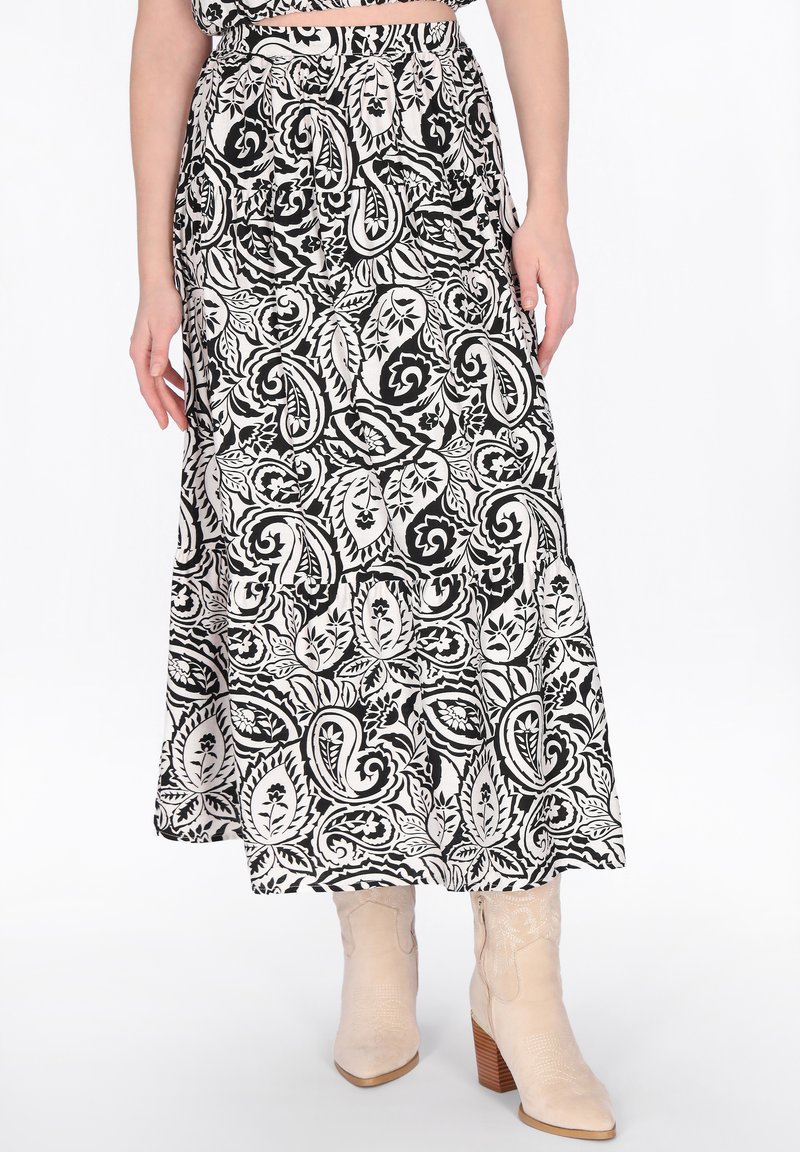Schwarz-weißer Paisley-Muster-Midirock, getragen mit beigefarbenen, Absatzstiefeln, an einer Person stehend vor weißem Hintergrund.