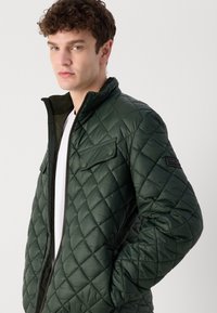 Veste verte matelassée avec un col montant, fermeture éclair et détails de poches à pression. Présente une surface texturée et un design ajusté.