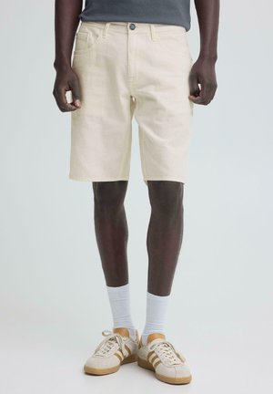Man wearing beige knee-length shorts, white crew socks, and off-white sneakers with brown stripes, standing on a plain light background.