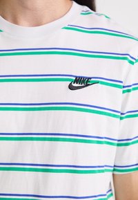 Bílá bavlněná tričko s modrými a zelenými horizontálními pruhy. Na levé části hrudi se nachází černě vyšitá značka Nike.